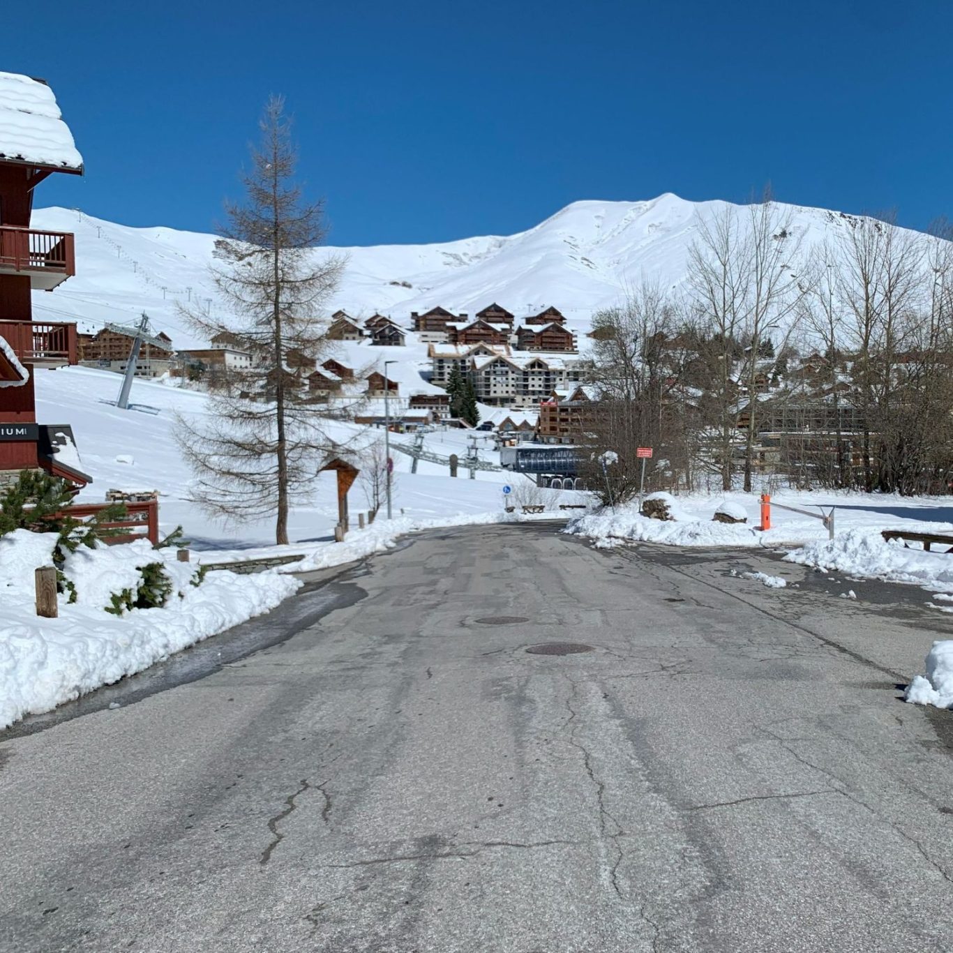 Une route enneigée avec des chalets en bois sous un ciel bleu. Montagnes en arrière-plan.