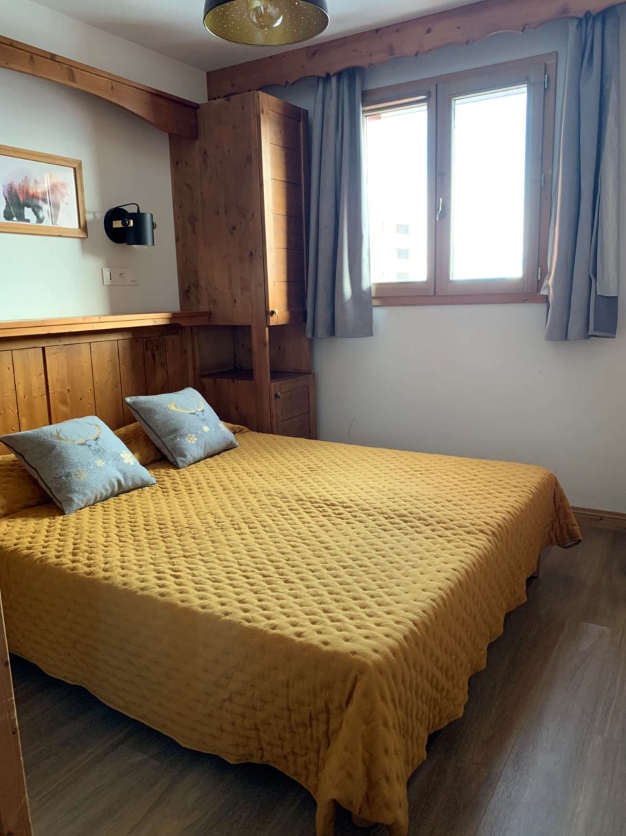Chambre avec lit double, couverture jaune et fenêtre apportant de la lumière naturelle.